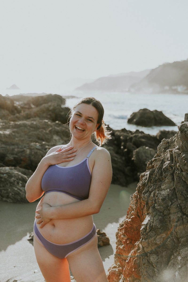 Woman in a lilac bra standing on a rocky beach with ocean in the background